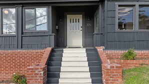 Uma casa de dois andares com fachada cinza escura, porta frontal branca e escadaria de tijolos que dá acesso à entrada.