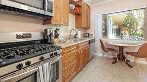 A kitchen with wooden cabinets, a gas stove, a microwave, and a dining area with a table and chairs. - PB Local Living Beach Access Less Than a Block (San Diego)