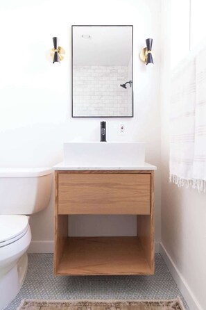 A bathroom with a white toilet, a wooden vanity with a mirror above it, and a small shelf below. - Step Into Style: Walkable Living in South Park (San Diego)