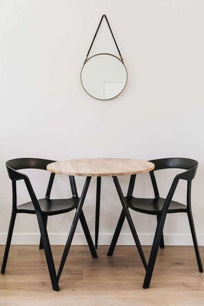 A round wooden table with black metal legs, flanked by two black chairs, against a plain white wall with a hanging circular mirror. - Step Into Style: Walkable Living in South Park (San Diego)