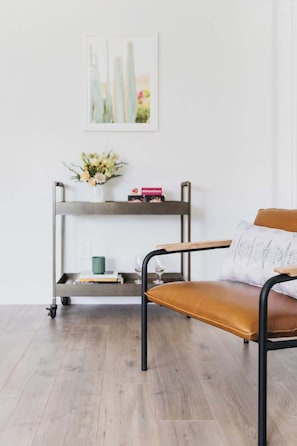 Een moderne kamer met een houten vloer, een bruine leren bank, een stoel met metalen frame, een metalen plank met een bloemenvaas, en een abstract schilderij in een lijst aan de muur.