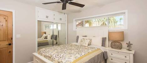 A bedroom with a bed, a nightstand, a ceiling fan, a window with a view, and a door.