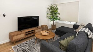 Ein modernes Wohnzimmer mit Flachbildfernseher, einer Holz-Unterhaltungseinheit, einem runden Couchtisch, einem grauen Sofa und einer großen Topfpflanze.