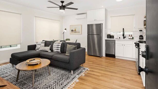 Eine moderne Wohnung mit grauem Sofa, einem kleinen runden Tisch, einer Küche mit weißen Schränken und Edelstahlgeräten sowie einem Deckenventilator.