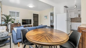 A modern living room with a round wooden dining table, blue sofa, and a flat-screen TV. - Quiet, Comfortable Sunset Cliffs Ocean Beach Apartment – Walk to Beach! (San Diego)