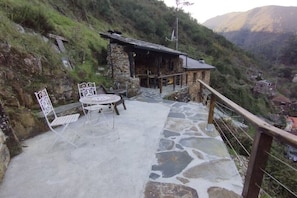 A stone patio with a table and chairs, a stone building, and a mountainous backdrop.