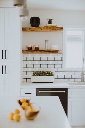 Cozinha moderna com armários brancos, painel de fundo preto e prateleira de madeira com livros e uma planta.