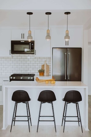 Cozinha moderna com ilha central, três banquetas pretas, eletrodomésticos de aço inoxidável e iluminação pendente.