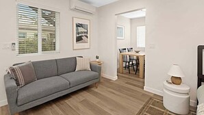 Ein modernes Wohnzimmer mit einem grauen Sofa, einem kleinen Beistelltisch mit einer Lampe und einem Essbereich mit einem Holztisch und Stühlen.