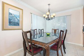 A dining room with a glass-top table, wooden chairs, a chandelier, a framed picture on the wall, and a window with blinds.
