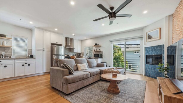 A modern kitchen with white cabinets, a grey sofa, a wooden coffee table, and a ceiling fan. - Stylish 2BD Peek-a-boo Ocean View High-End Stay (Solana Beach)