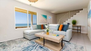 A modern living room with a white sofa, a coffee table, and a view of the ocean through large windows. - 4BR Home Sunset Cliffs Oceanfront - Cliffside View (San Diego)