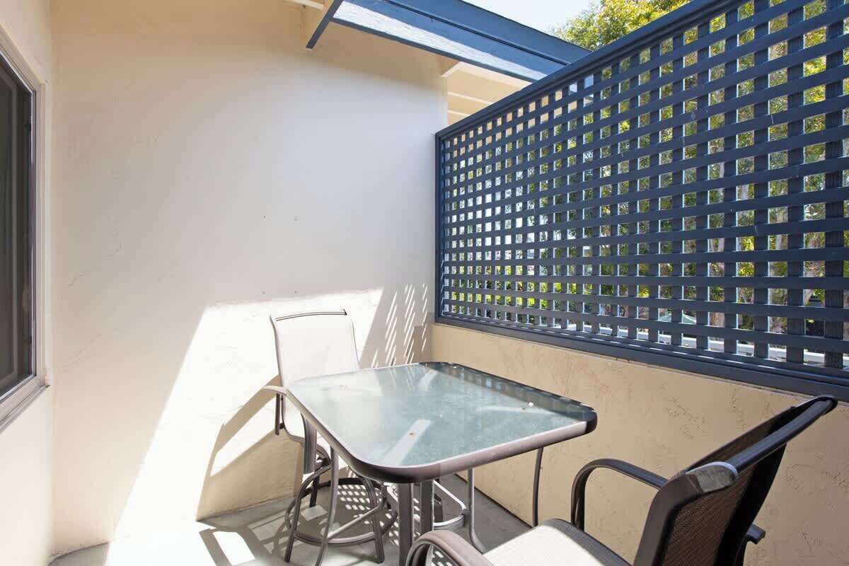 A small outdoor patio with a glass table and metal chairs, a blue metal fence, and a window on the left.