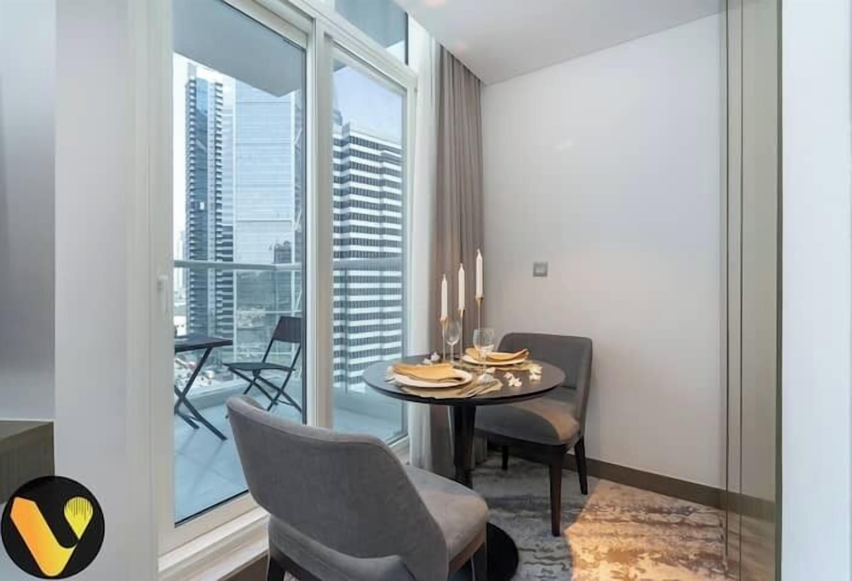 A modern hotel room with a large window, a round dining table set with plates, glasses, and candles, and a grey upholstered chair.