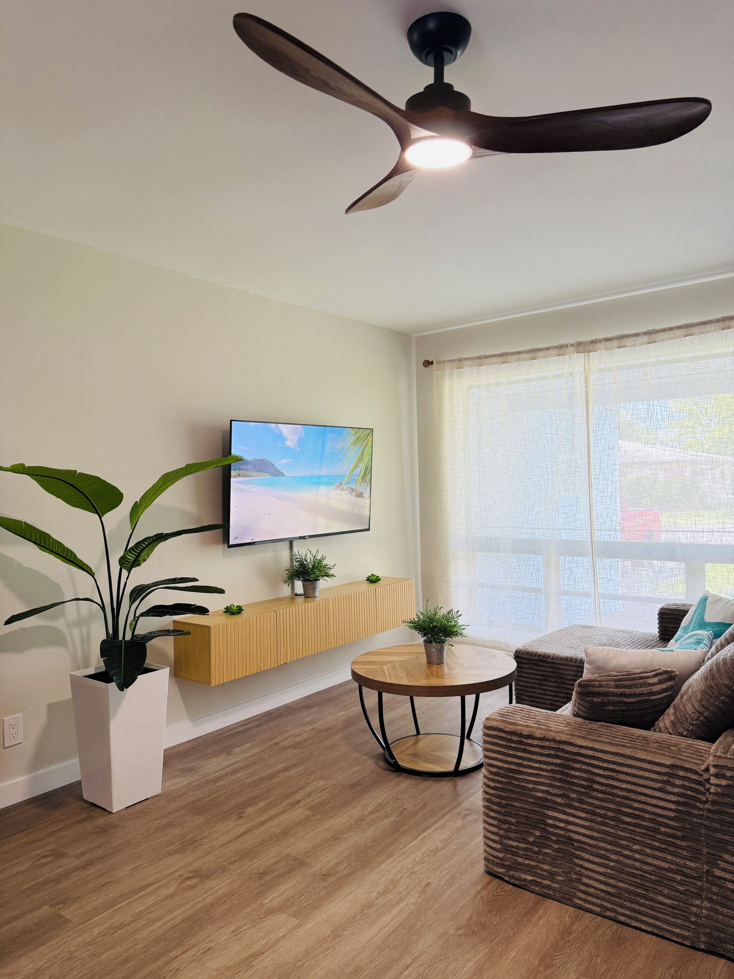 Ein modernes Wohnzimmer mit Deckenventilator, Flachbildfernseher, Couchtisch aus Holz, braunem Sofa und großem Fenster mit transparenten Vorhängen.