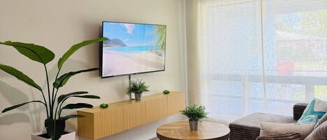 Ein modernes Wohnzimmer mit Deckenventilator, Flachbildfernseher, Couchtisch aus Holz, braunem Sofa und großem Fenster mit transparenten Vorhängen.