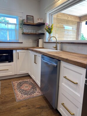 Una cocina con armarios blancos, lavavajillas de acero inoxidable, encimera de madera y ventana con vistas a zonas verdes.