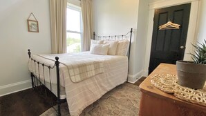 Quarto com cama, janela com cortinas, porta com cabide e uma pequena planta sobre uma mesa.