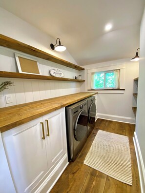 Lavandería con lavadora, repisas de madera, una ventana con cortinas y una lámpara.