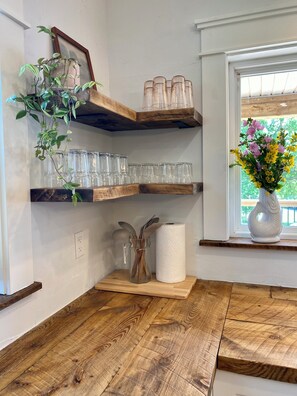 Una esquina de una habitación con estantes de madera que sostienen cristalería y un jarrón con flores.