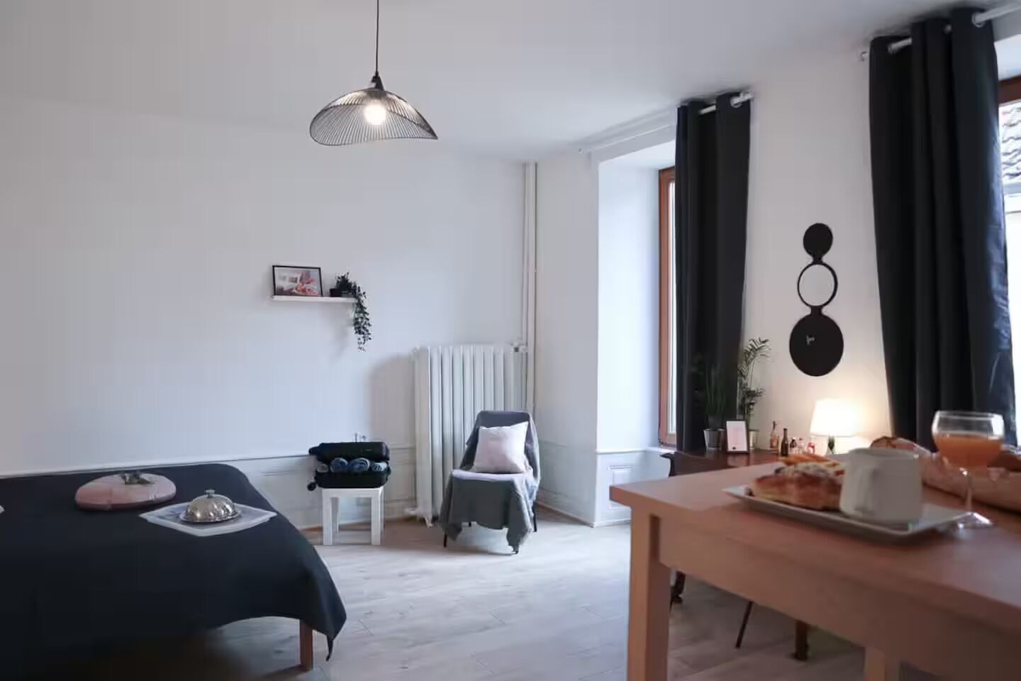 A modern hotel room with a bed, a dining table with breakfast, a chair, a radiator, and a hanging light fixture.