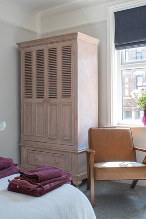 Une chambre dotée d’une grande armoire, d’une chaise et d’une fenêtre donnant sur des immeubles.