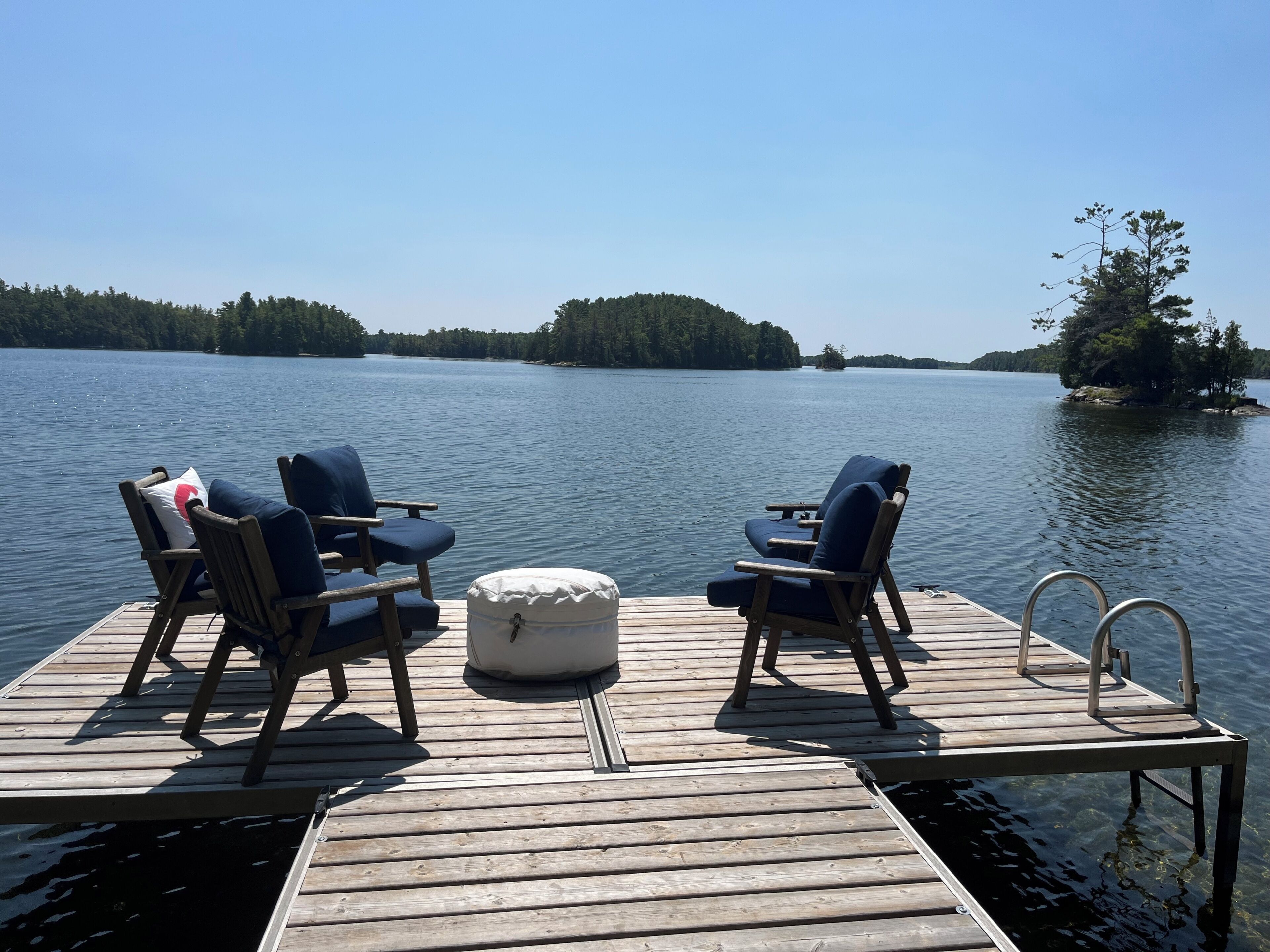 Un muelle de madera con dos sillones de jardín y un reposapiés acolchado, con vistas a un lago tranquilo y una isla boscosa en la distancia.