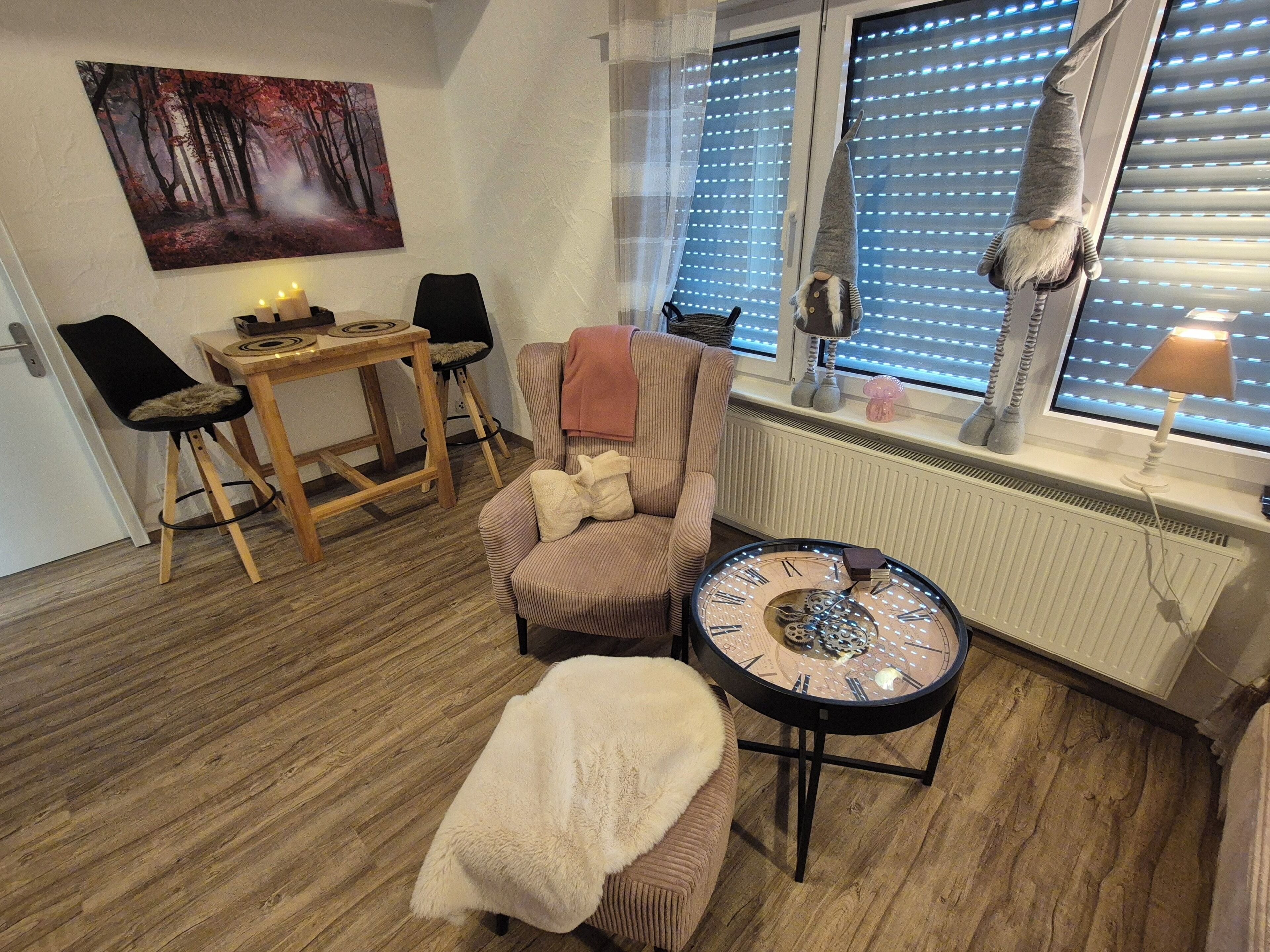 A cozy room with a wooden floor, a round table with a clock, a chair, a sofa, a window with blinds, and a painting on the wall.