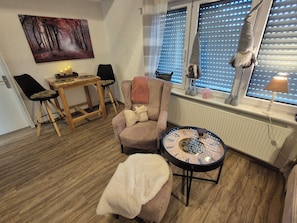 A cozy room with a wooden floor, a round table with a clock, a chair, a sofa, a window with blinds, and a painting on the wall. - Cozy vacation apartment in Medebach Dreislar 15 km from Winterberg in the Sauerland (Medebach)