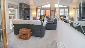 A spacious living room with a fireplace, grey sofas, and a large window overlooking a garden. - Waters Edge Lodge (Windermere)