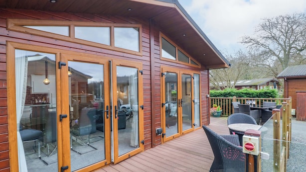 A wooden building with large glass doors and a deck area with outdoor furniture. - Waters Edge Lodge (Windermere)