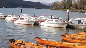 A marina with several small boats docked, a wooden pier, and a forested hill in the background. - Waters Edge Lodge (Windermere)