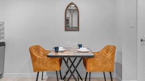 Un comedor con una mesa preparada para dos personas, dos sillas de color mostaza, un espejo en la pared y una zona de cocina a la izquierda.