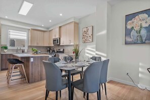 A modern kitchen with wooden cabinets, a central island, and a dining area with grey chairs. - Stylish 3BR Townhouse Near U of A (Edmonton)
