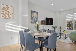 A dining area with a round table set for four, surrounded by grey chairs. There are two paintings on the wall, a potted plant, and a TV mounted above the table.