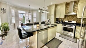 A modern kitchen with a central island, stainless steel appliances, and a dining area with black chairs. - Spacious 4BR Home with Private Yard & Fast WiFi (Brampton)