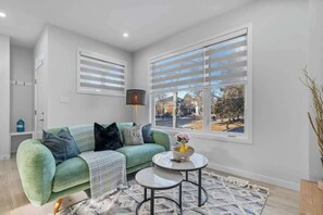 A living room with a green sofa, a small round table, and a large window with blinds.