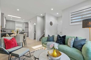 A modern living room with a green sofa, a grey armchair, and a coffee table with a vase. A kitchen is visible in the background with white cabinets and a dining area.