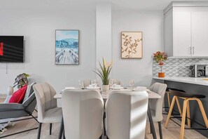 A modern dining area with a white table set for six, surrounded by beige chairs. There are two framed artworks on the wall, a potted plant on the table, and a kitchen area with a microwave and coffee maker visible.
