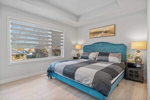 A bedroom with a large bed, a window with blinds, a painting on the wall, and a nightstand with a lamp.