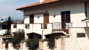 Ein zweistöckiges Gebäude mit einem Ziegeldach, weißen Wänden und einem Steinunterbau. Es gibt Fenster mit Fensterläden, einen Balkon mit Metallgeländer und eine kleine Mauer im Vordergrund.