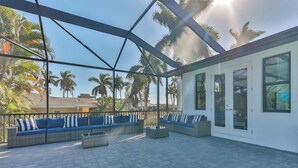 A covered outdoor seating area with a modern design, featuring a sofa set and a small table, overlooking palm trees and a residential area.