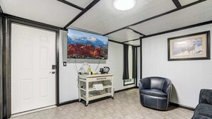 Una habitación con una puerta blanca, un televisor montado mostrando un paisaje de montaña, un escritorio blanco con cafetera, un sillón de cuero negro y un cuadro en la pared.