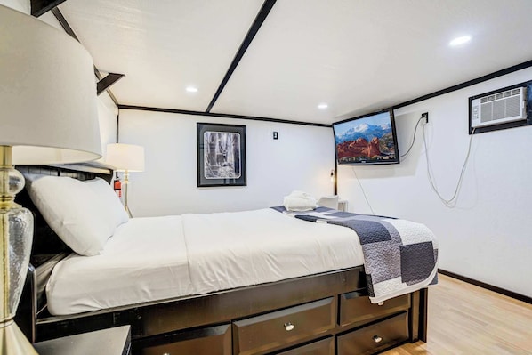 A neatly arranged bedroom with a bed, bedside lamp, a framed picture, a television, and a wall-mounted air conditioning unit.