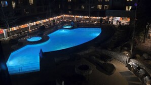 Zona de piscina por la noche con varias piscinas, asientos de piedra y un patio bien iluminado.