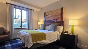 Habitación de hotel con una cama grande, dos lámparas de noche, una silla, un cuadro en la pared y una ventana con cortinas.