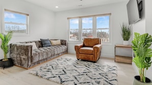 A modern living room with a large window, a ceiling fan, a brown leather armchair, and a grey sofa with multiple cushions. - Cozy Home in Newton (Newton)