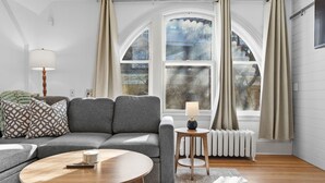 Uma sala de estar com um sofá cinzento, uma mesa de centro redonda de madeira, uma pequena mesa lateral com abajur e uma grande janela em arco com cortinas.