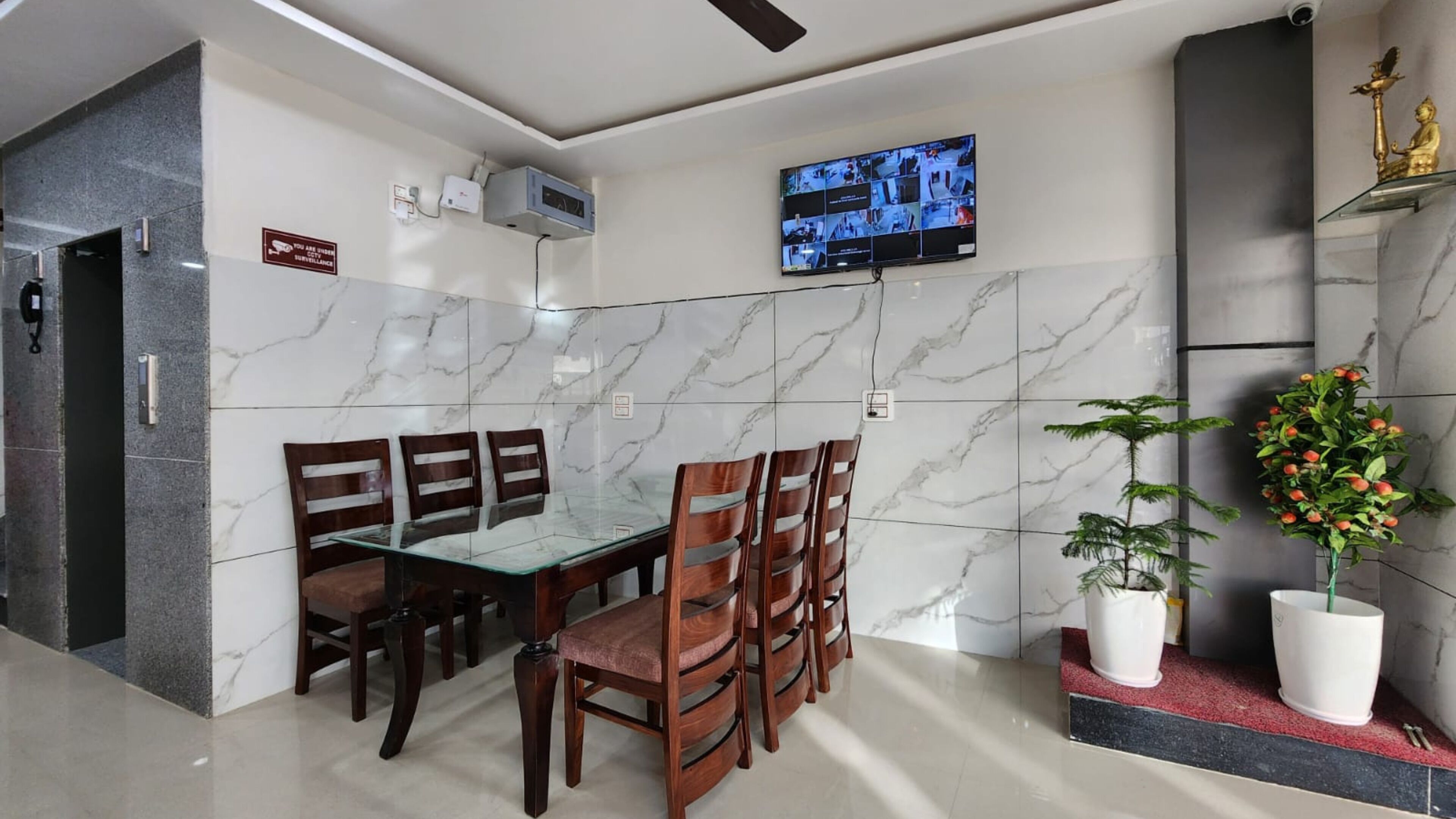 A modern dining area with a glass table and wooden chairs, a mounted TV displaying multiple camera feeds, and a decorative Buddha statue on a shelf.
