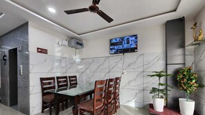 A modern dining area with a glass table and wooden chairs, a ceiling fan, a mounted TV, and potted plants. - Hotel The Orchid Plaza (Samba)